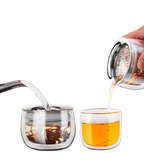 Double-wall glass infuser pot brewing loose-leaf tea beside a separate glass drinking cup